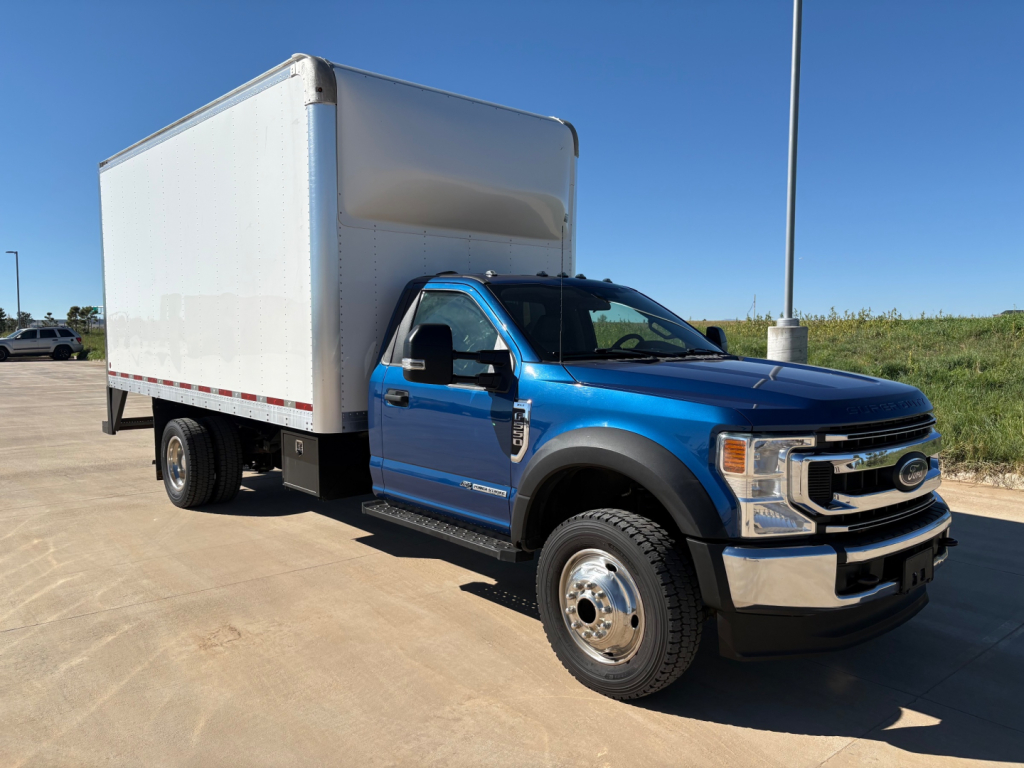 Box truck with a blue Ford F-550 cab and a large white cargo box, front three-quarter view, daytime clear sky.