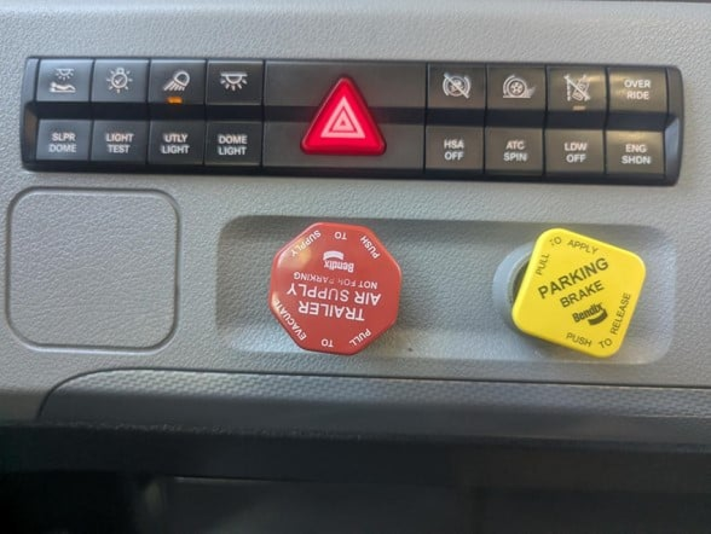 Dashboard controls of a Freightliner Cascadia 126 showing hazard light button, parking brake yellow knob, red power knob, and various switch labels.