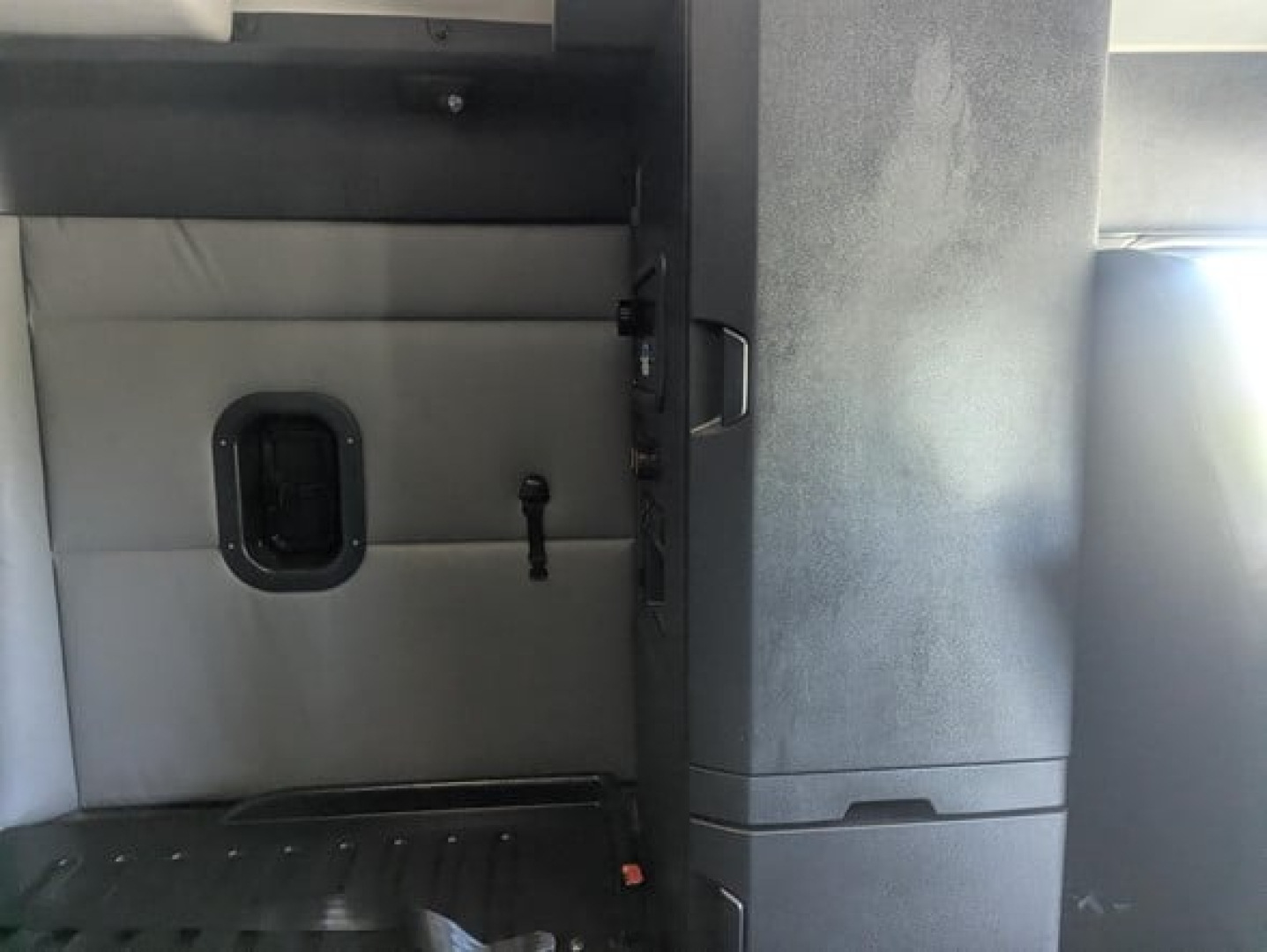 Freightliner Cascadia cab interior showing partition wall with a small rectangular window and a door latch; light gray upholstery and black trim.