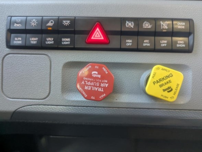 Dashboard controls of a Freightliner Cascadia 126 showing hazard light button, parking brake yellow knob, red power knob, and various switch labels.