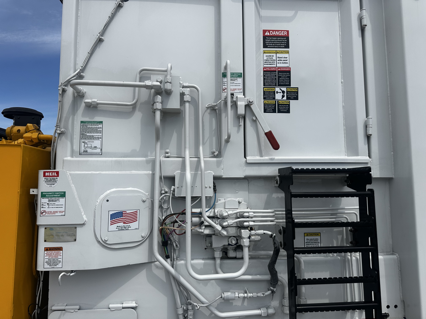 Truck rear door with hydraulic lines and safety stickers. Visible text includes DANGER label, various warning tags, small American flag sticker, and maintenance labels near hoses and control levers.