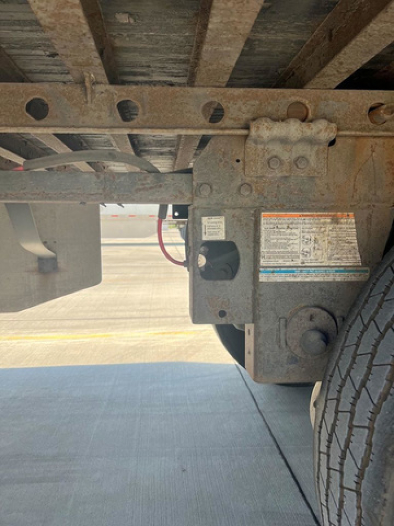 Rear undercarriage of a utility van showing a metal frame, suspension components, and a tire; visible are a rectangular metal block with bolts and a label, plus a small electrical/air line sheath.