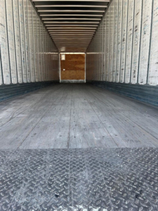 Trailer interior showing empty cargo space with metal side walls and wooden rear door, diamond plate threshold, E-tracks along sides.