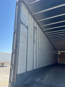 Inside of an empty trailer with vertical wall panels and door posts. Visible markings include '53 X  ...' on the wall and a partially legible '46' near the door jamb, with metal ribbed ceiling and corner supports.