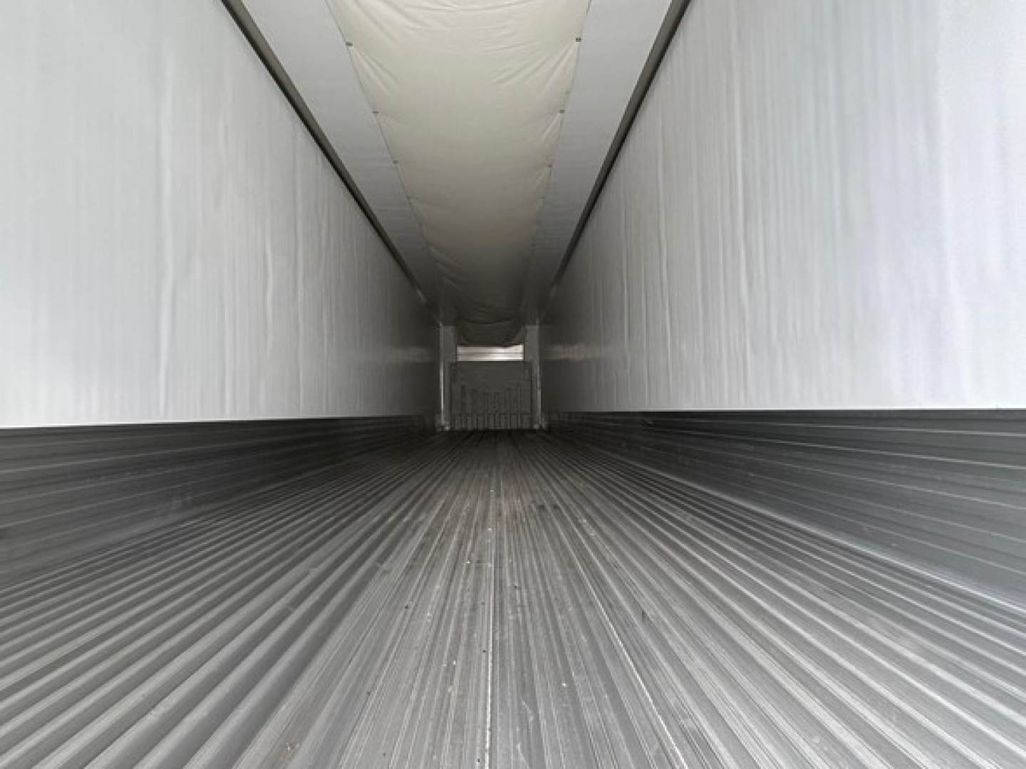 Trailer interior showing long corrugated metal floor and white sidewalls with ceiling; viewed from front toward the rear with a hatch at far end.