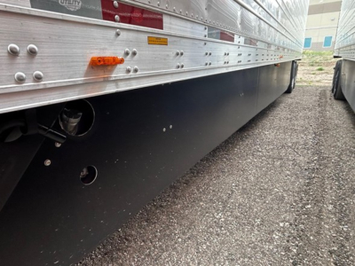 Trailer side with lower black mud flap area showing riveted aluminum siding and wheel area; small orange reflector and adhesive labels visible.
