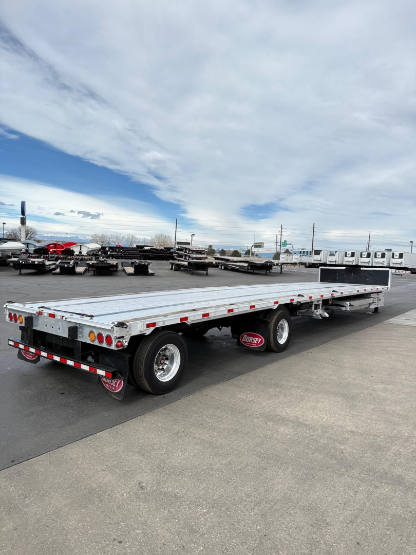 Long flatbed trailer with drop deck showing dual rear axles and reflective tape, outdoor lot with other trailers in background.