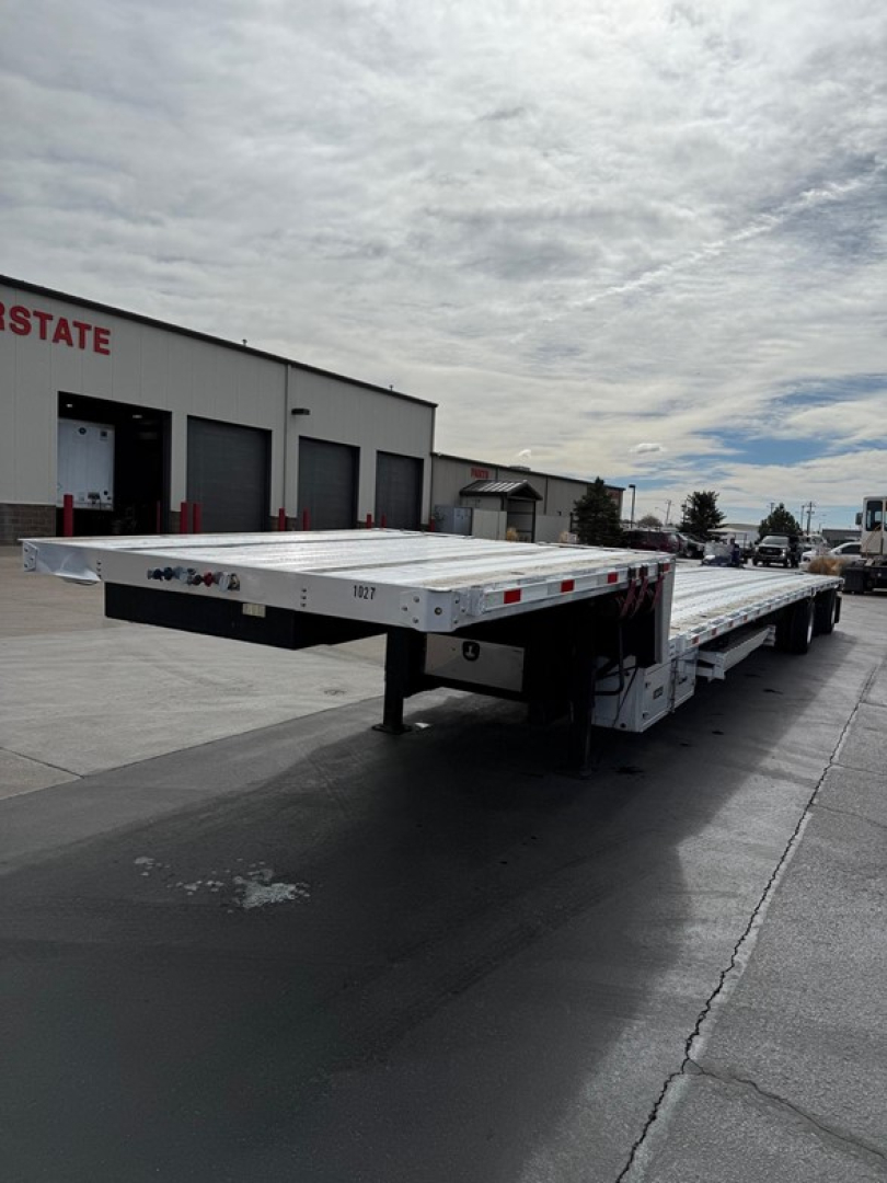 Flatbed trailer in side view showing a Dorsey combo drop deck with reflective tape and aluminum deck, light gray color, at a dealership lot.