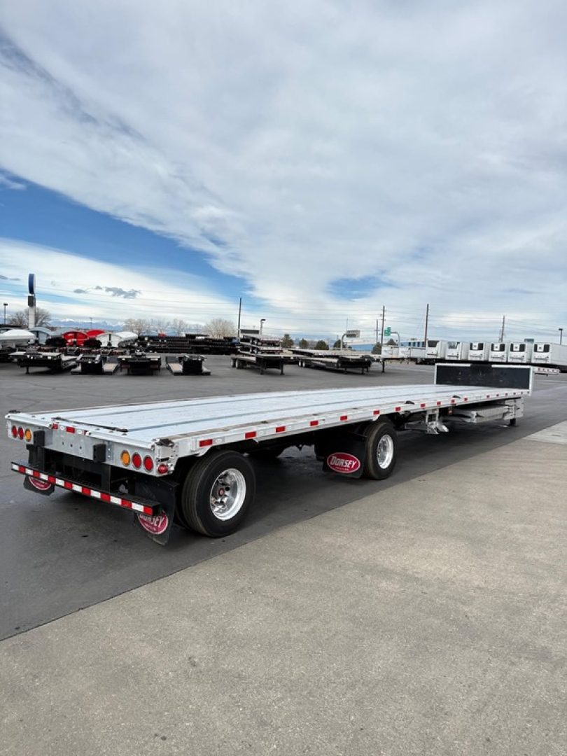 Heavy multi-axle drop deck trailer in an outdoor lot, aluminum flatbed with dual rear axles, reflective tape, and Dorsey branding on mud flaps in the distance.
