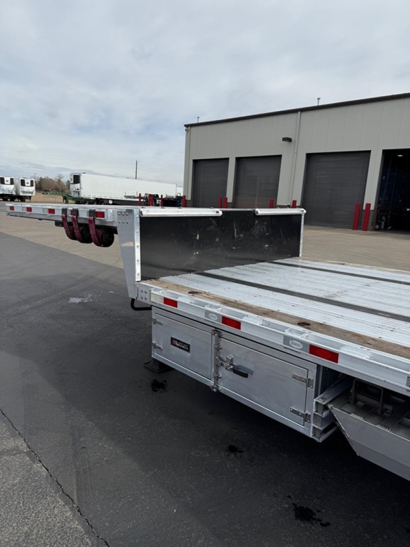 Drop deck trailer with dual-level loading platform and rear storage compartments showing aluminum deck and caution markings.