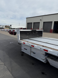Aluminum drop deck trailer with extendable ramp deck and open flatbed area, rear taillights visible and a warehouse in background.