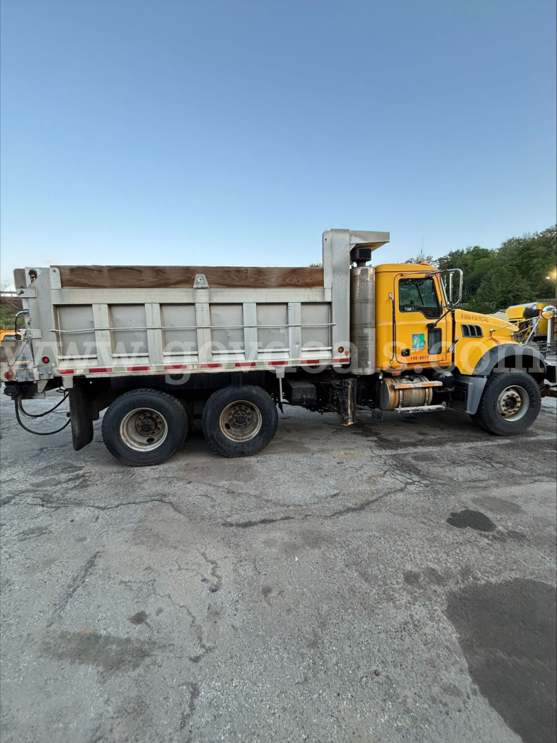 Truck side view showing a yellow Mack GU713 dump truck with silver dump bed and wooden side rails.