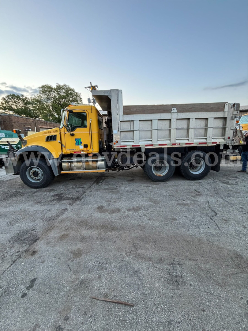 Dump truck side view showing yellow Mack GU713 with gray dump bed and red-and-white reflective tape.