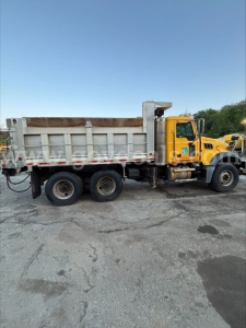 Truck side view showing a yellow Mack GU713 dump truck with silver dump bed and wooden side rails.