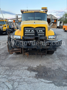 Mack GU713 dump truck front view showing large chrome grille with MACK logo, heavy bumper, headlights, and snow plow attachment at the front; yellow cab with black bumper area.