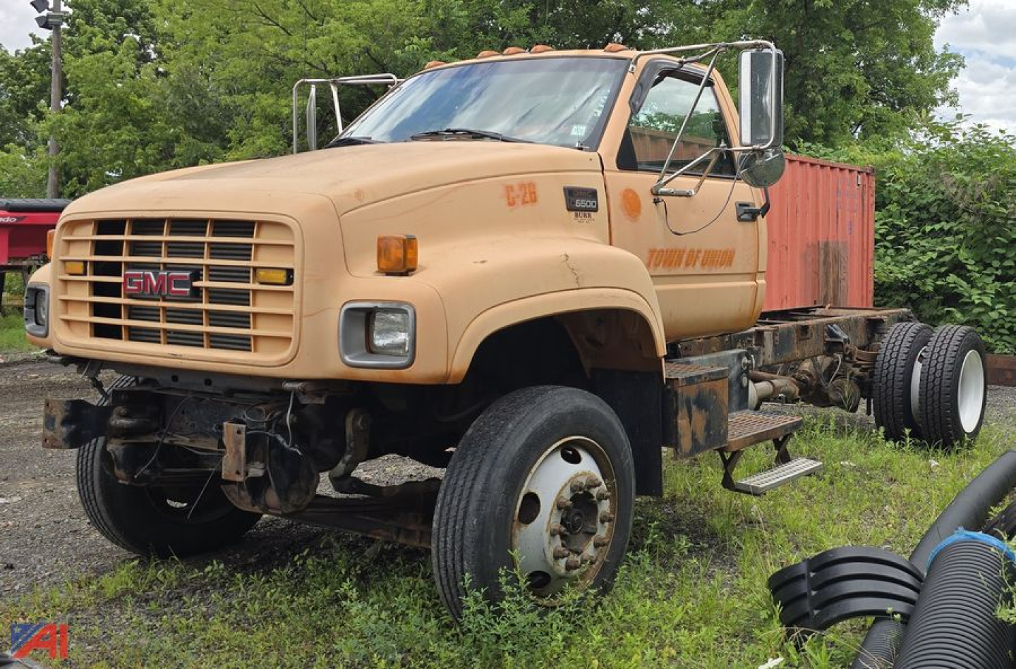 GMC C7500 cab and chassis truck showing front grille with GMC emblem and beige paint, missing front bumper, dual rear axles, and off-road tires.
