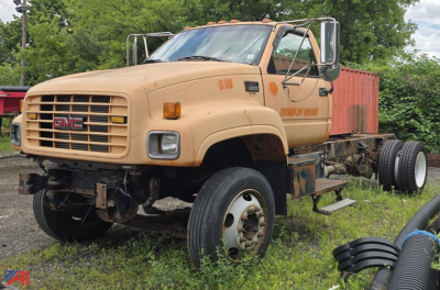 GMC C7500 cab and chassis truck showing front grille with GMC emblem and beige paint, missing front bumper, dual rear axles, and off-road tires.