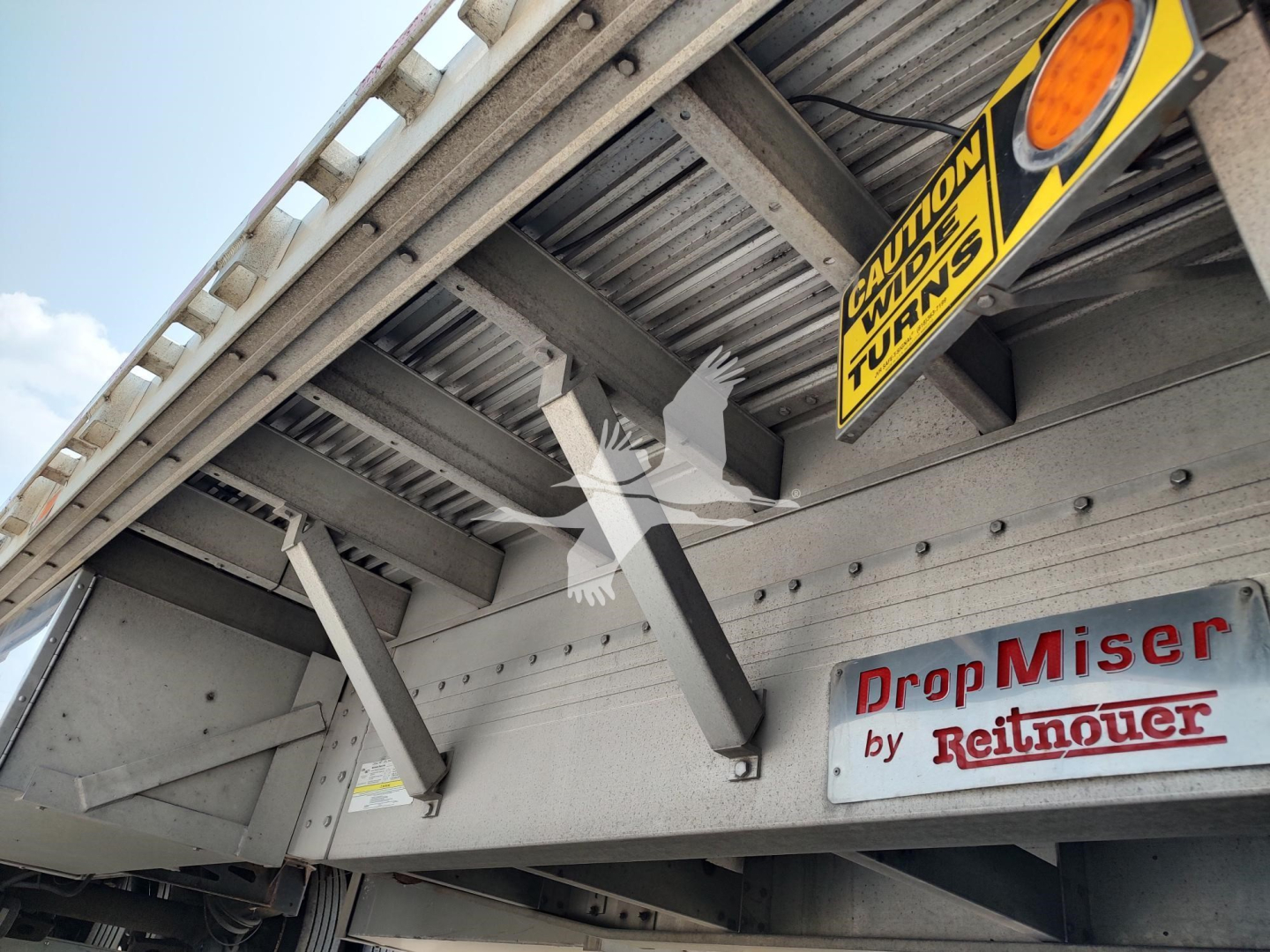 Drop deck trailer undercarriage with 'Drop Miser by Reitnouer' emblem and warning signs; visible structural beams and a yellow caution sign.