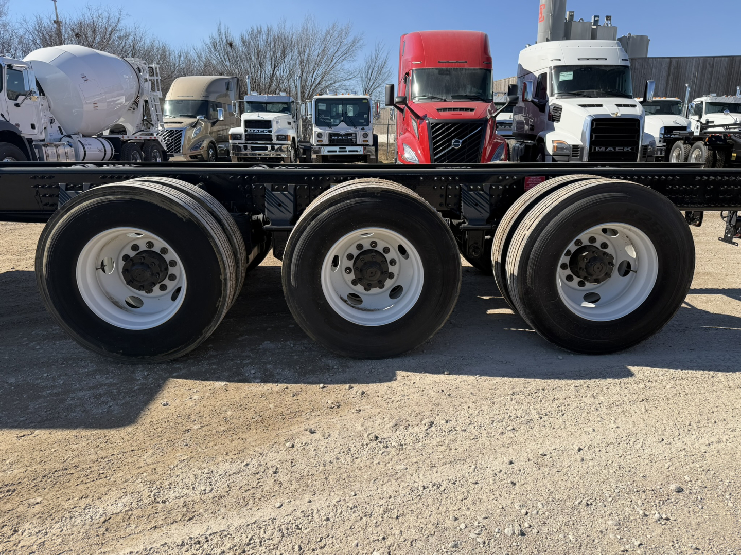 Volvo VHD86B300 cab and chassis wheelset view showing triple rear axle wheels and surrounding trucks in a lot.