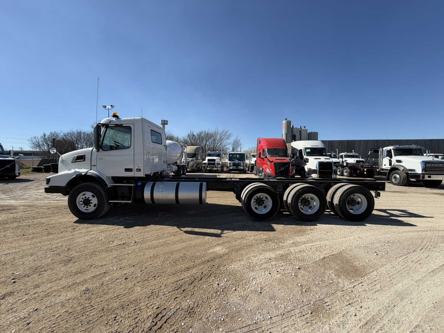 Volvo VHD86B300 cab and chassis truck in a dealership lot, side view, showing white cab, long chassis with multiple axles and fuel tank.