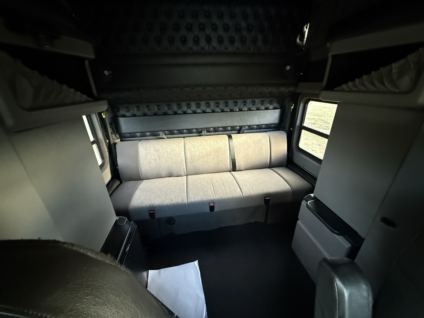 Kenworth W900L cab interior showing sleeper area with grey fabric seat, storage compartments, and windows on both sides.