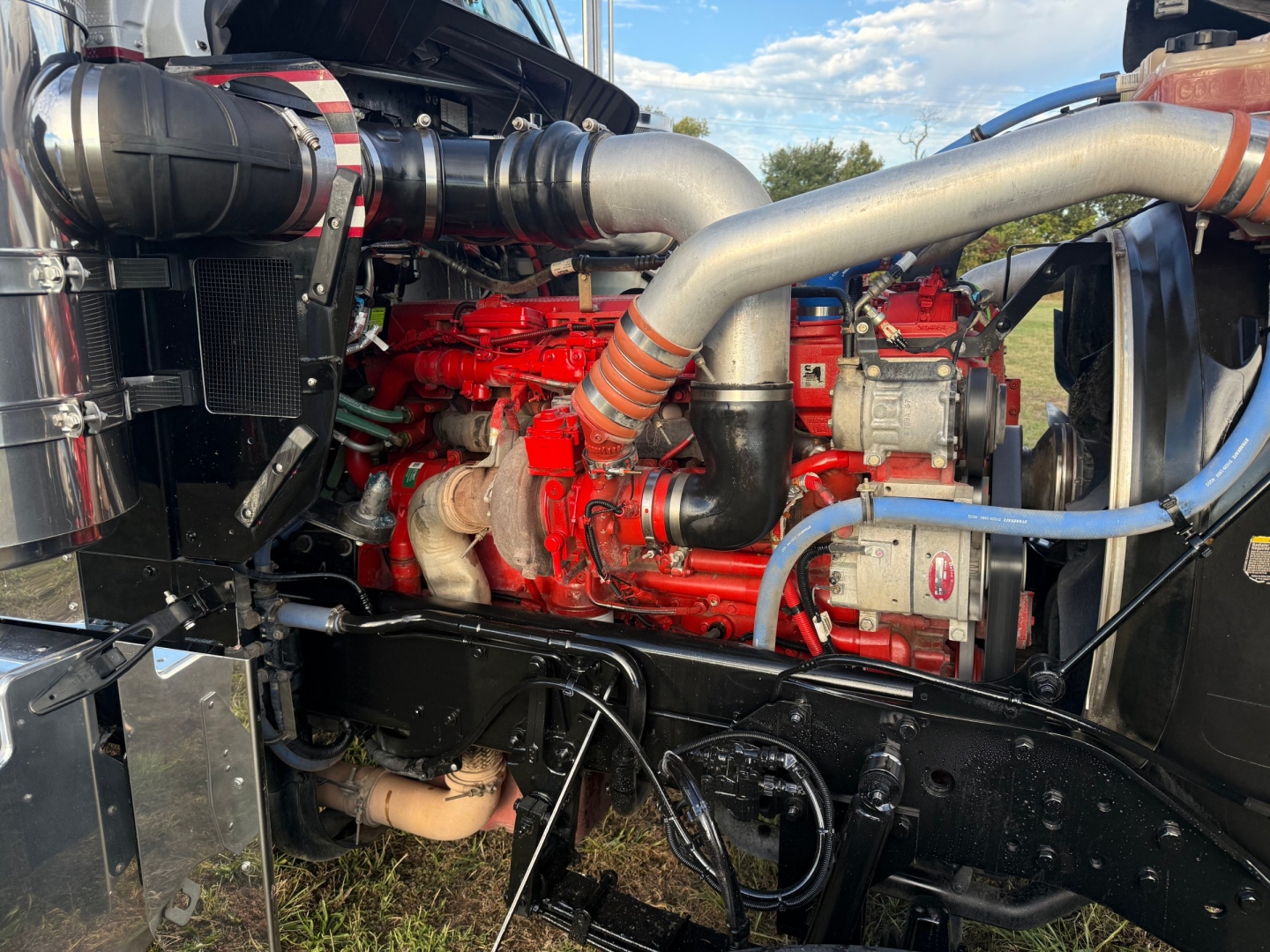 Kenworth W900L engine bay showing red engine with large silver exhaust manifolds, blue and red hoses, and black radiator area.