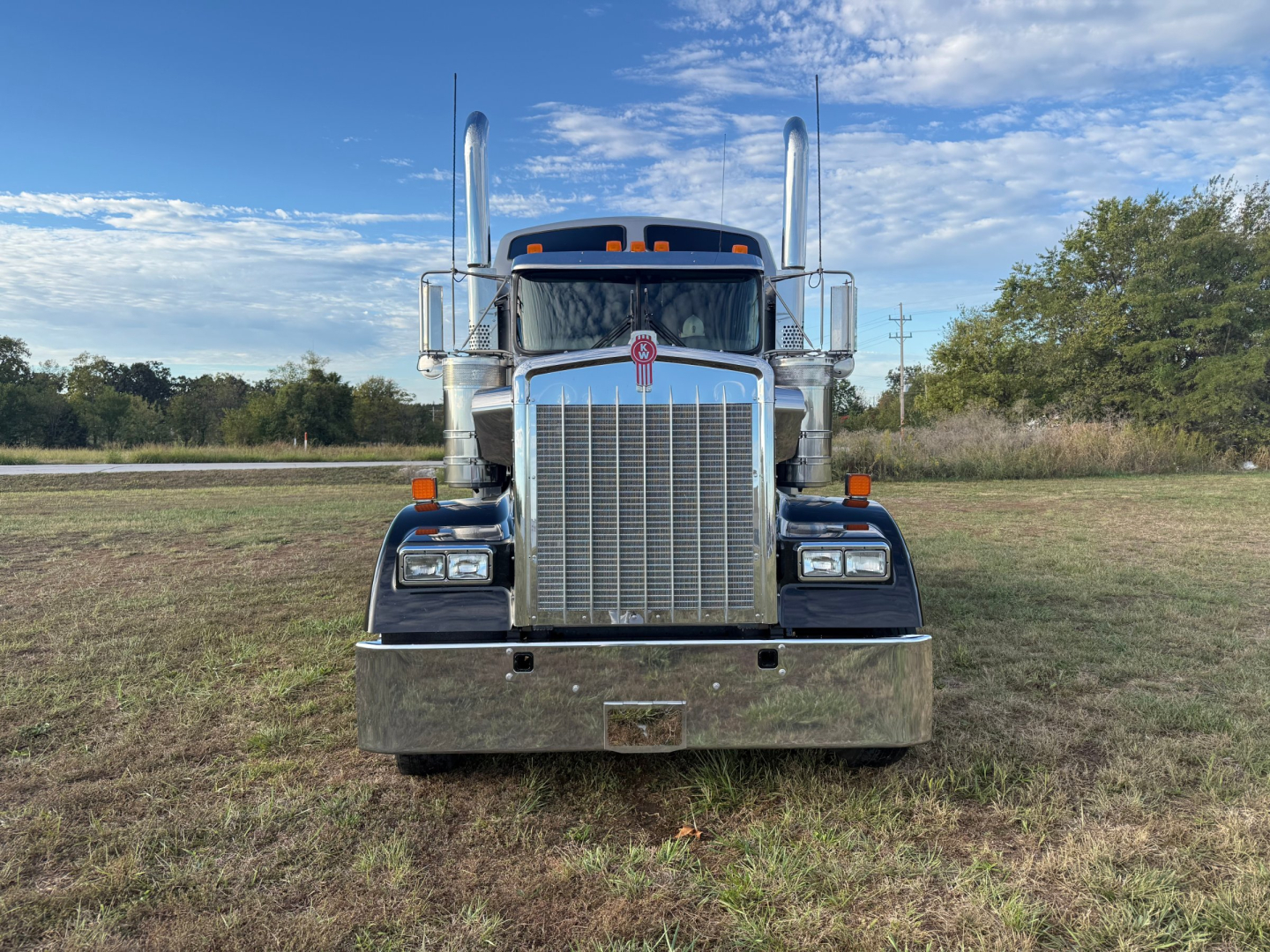 Kenworth W900L truck front view showing chrome grille, large bumper, dual chrome exhaust stacks, and headlights in a grassy lot.