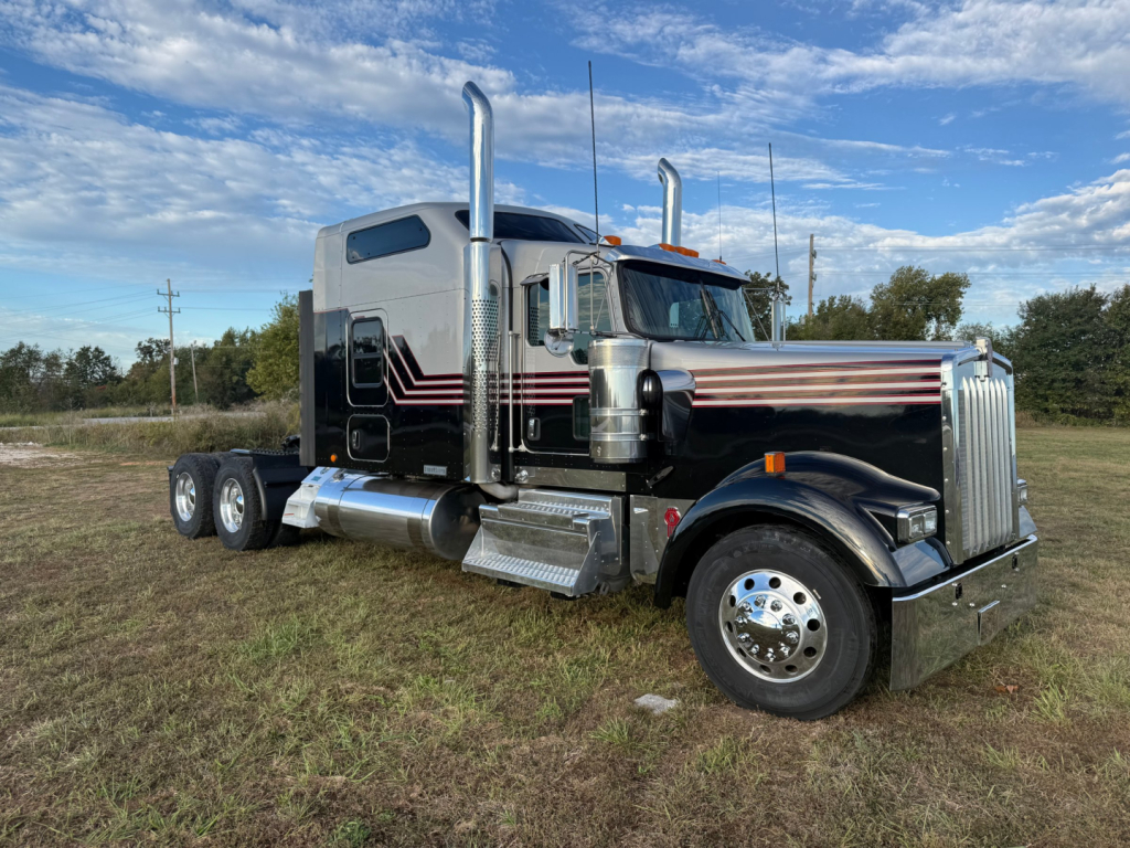 Kenworth W900L conventional sleeper truck in a grassy lot with chrome exhaust stacks and polished chrome bumper, black with red pinstriping.