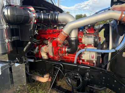 Kenworth W900L engine bay showing red engine with large silver exhaust manifolds, blue and red hoses, and black radiator area.