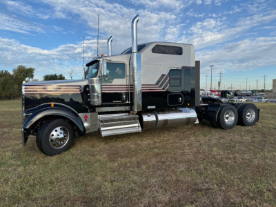 Kenworth W900L sleeper tractor photographed in a field. Side view shows chrome exhaust stacks, black and silver paint with red stripes, chrome fuel tanks and steps. Six-wheel drive rear with third axle visible.