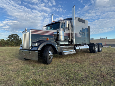 Kenworth W900L conventional sleeper truck in a field, chrome exhaust stacks, large grille, blue-gray with white and red pinstriping, dual rear axles.