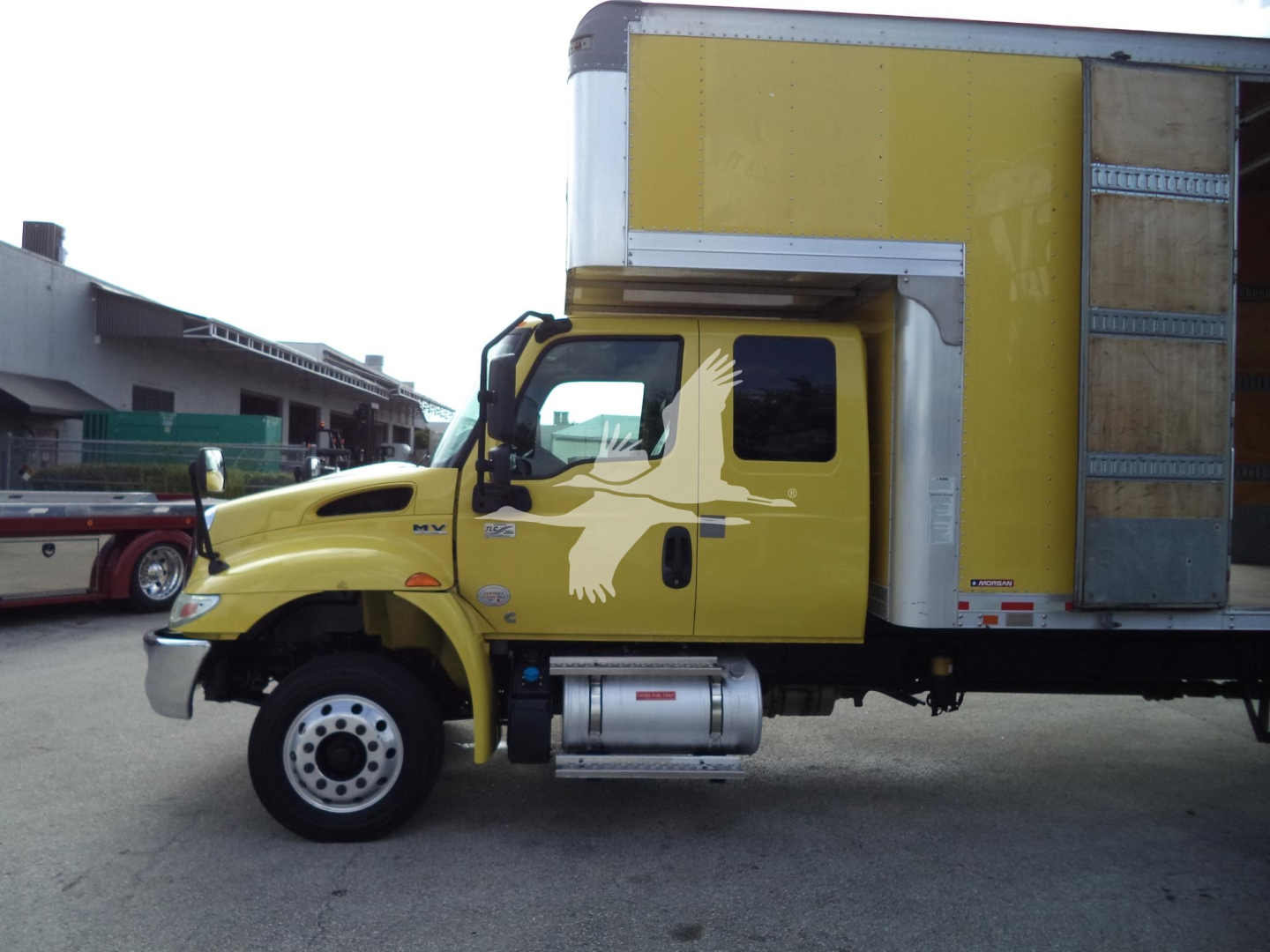 Truck cab and cargo box of a yellow 2020 International MV moving van, side view showing front wheel, fuel tank, and cargo box with wooden panel door.