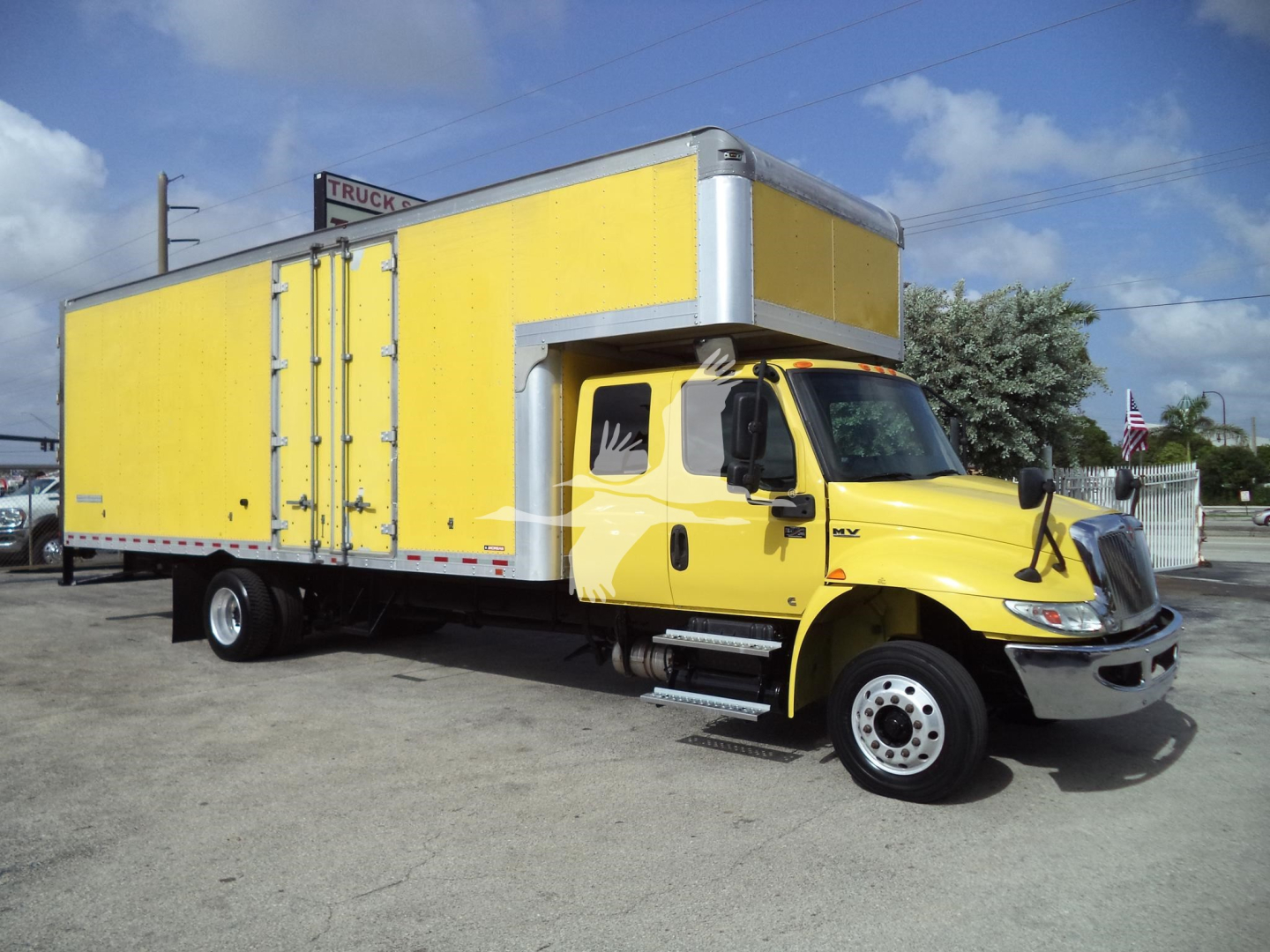 Yellow International MV moving van truck with large cargo box and chrome exhaust stack, side view showing cab and cargo area.