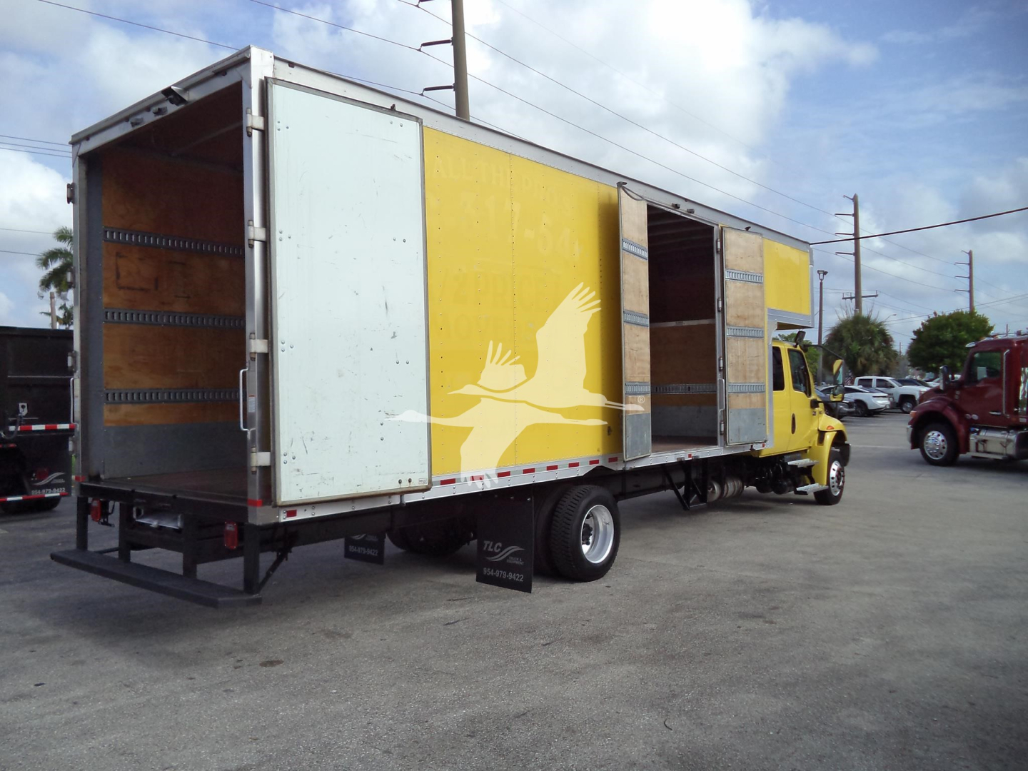 Moving van with yellow and white cargo box, side door open, showing interior walls and wooden floor panels. A small yellow cab is attached. Clear sky with some clouds.