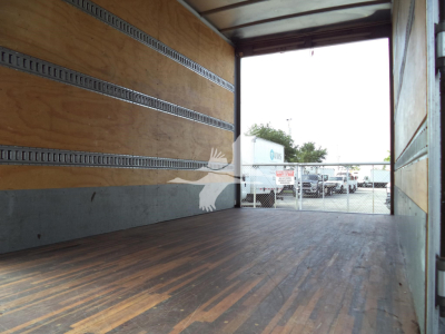 Interior of a cargo box with wooden floor and side rails; open back with chain-link fence and parked trucks outside.