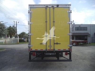 Rear view of a yellow box truck cargo trailer with double doors and metal latches, showing a white bird logo on the doors and a black bumper; surroundings include a building and parked equipment.