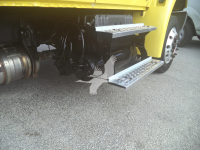 Truck side view with metal steps under yellow cab showing drivetrain components and wheel.