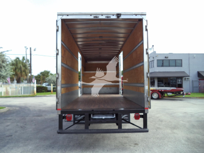 Trailer truck bed interior showing empty cargo area with wooden side walls and metal rails. Rear doors open, empty cargo floor, dealership lot in background.