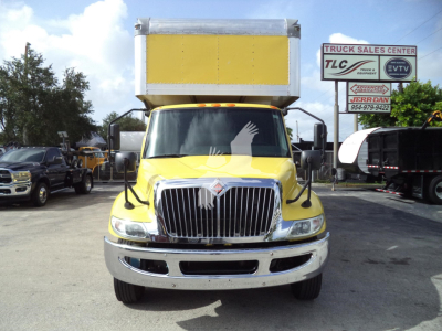 Front of a yellow 2020 International MV moving van with a tall box on top, large chrome grille, headlights, side mirrors, and a dealership sign in the background.