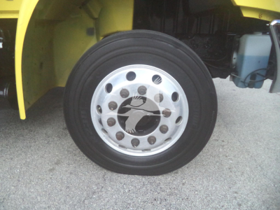 Wheel close-up on a yellow International MV truck showing a polished aluminum rim with multiple lug nuts and a tire; part of the chassis and coolant reservoir visible in background.