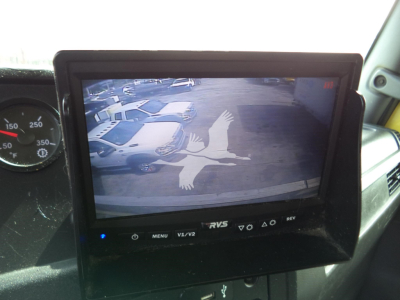 In-cab monitor displaying a bird logo on a rear-view camera screen; surrounding dashboard with gauge cluster visible.