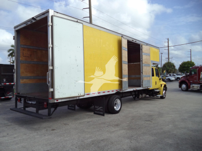 Moving van with yellow and white cargo box, side door open, showing interior walls and wooden floor panels. A small yellow cab is attached. Clear sky with some clouds.