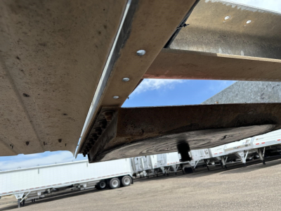 Trailer undercarriage showing metal frame and a black rubber bumper/edge with rivets; ground with other trailers in background.