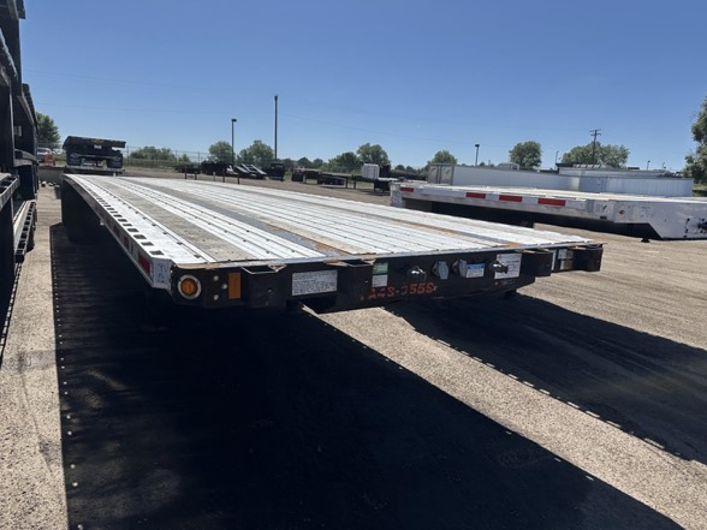 Flatbed trailer in a lot showing a long aluminum deck with rear lighting and reflective tape; visible text includes license plate area and small labels along the rear.