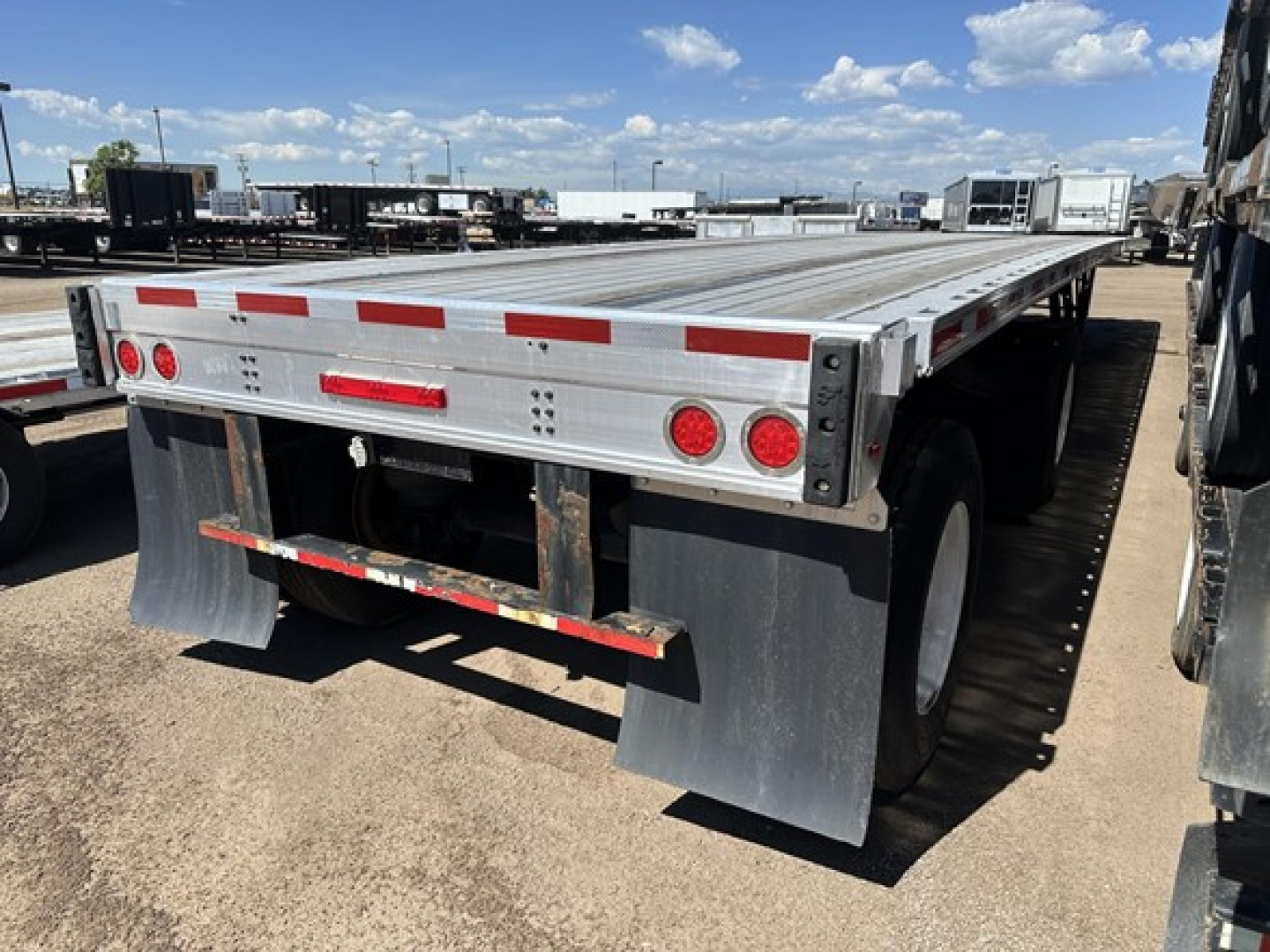 Flatbed trailer rear view showing dual rear tires, mud flaps, and reflective safety markings on aluminum deck with red lights and brake lights.