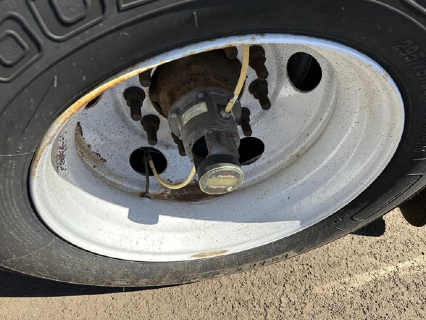 Wheel hub with tire showing valve stem and lug nuts; rim and brake drum visible in a trailer axle, light rust around edges.