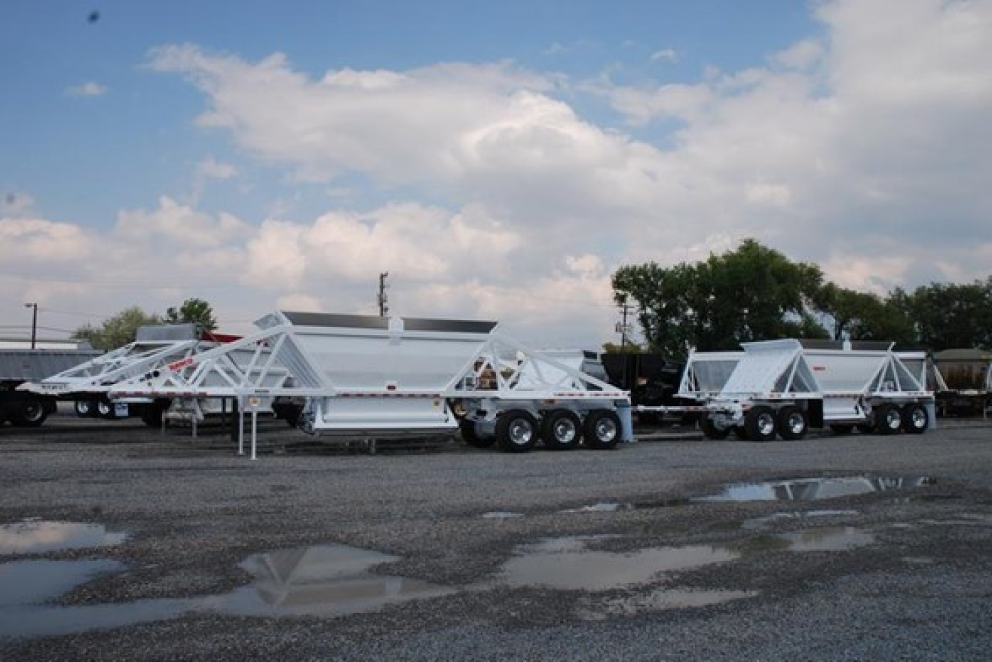 Lightweight bottom dump trailers lined up outdoors, white paint, multiple axles, blue sky with clouds.