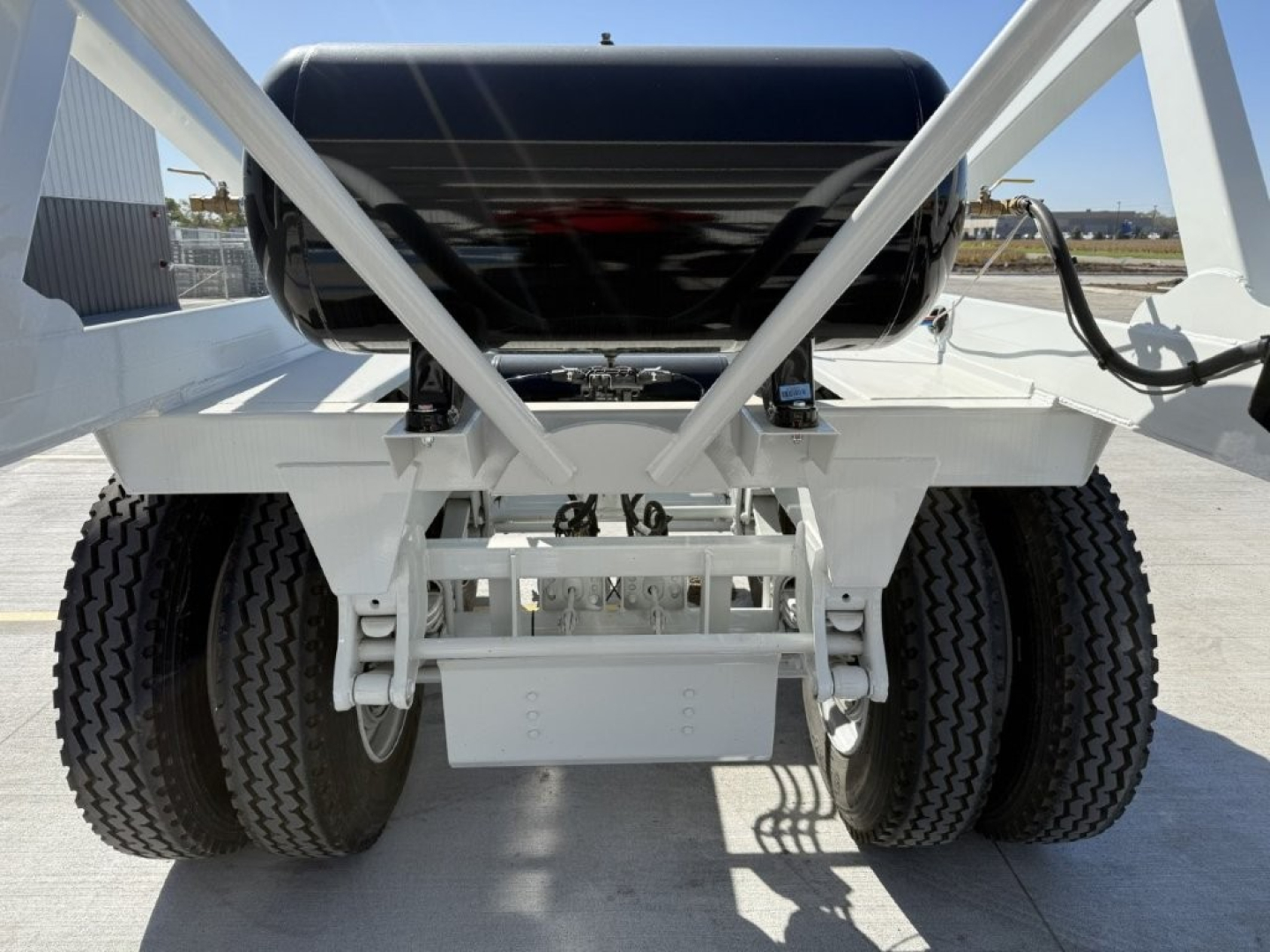 Dump trailer undercarriage showing dual tandem axles and suspension with spare tire rack, white frame, black cylindrical tank, and labeling not visible.