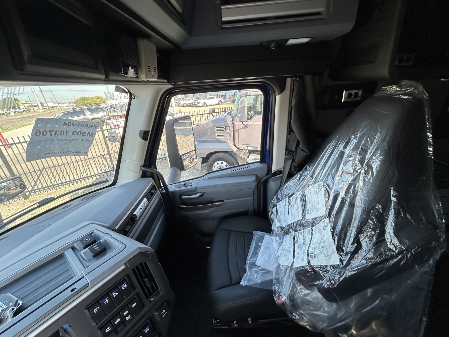 Cab interior of a 2027 Mack PR64T76H showing driver seat wrapped in plastic and dashboard controls visible.