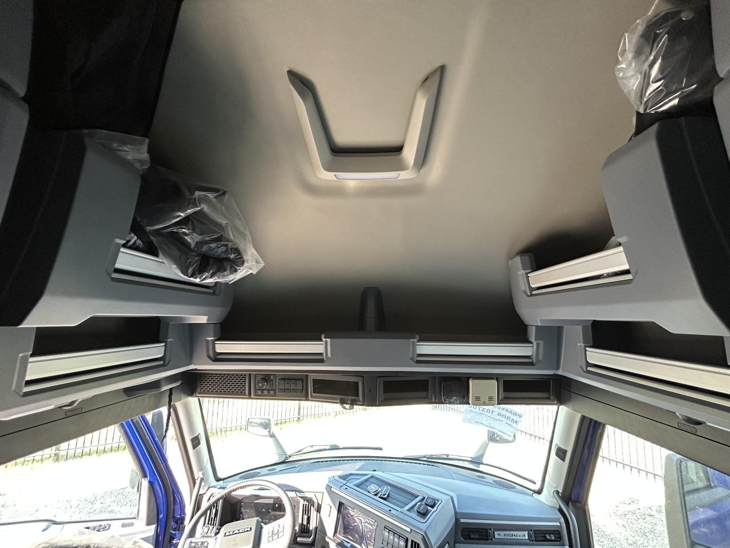 Mack truck interior cab view from above showing overhead storage and dashboard with steering wheel and center console, interior plastic-wrapped components.